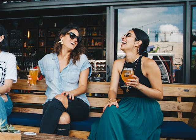 two people laughing and drinking beer on a restaurant bench